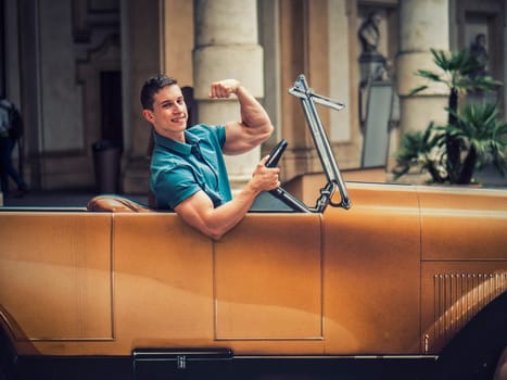 A young man is sitting in a fake vintage car, doing a bodybuilding bicep pose and smiling to the camera