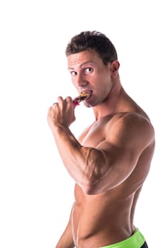 Handsome muscular shirtless man eating protein bar, isolated on white in studio, looking at camera