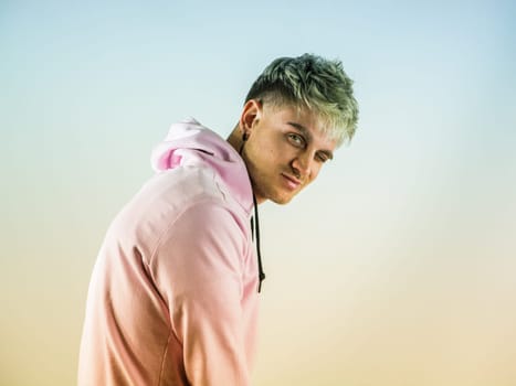 Attractive blond young man with blue eyes winking, wearing pink sweater, in studio shot on neutral background