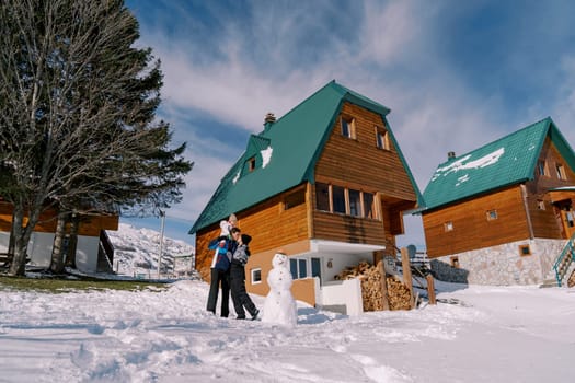 Mom kisses dad with little girl on his shoulders standing next to snowman near wooden chalet. High quality photo