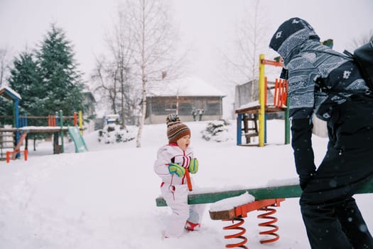 Mom and little girl swing on a snow-covered swing-balancer at the playground. High quality photo