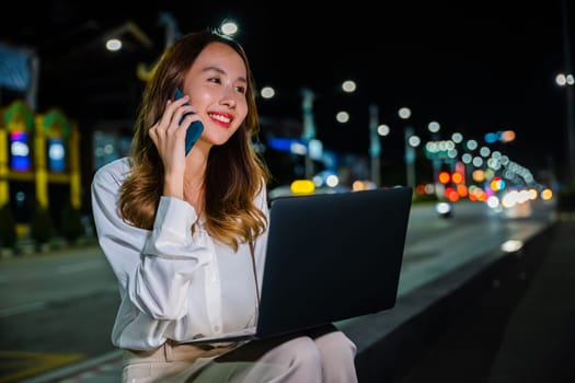 In the heart of the city at night, a woman is multitasking on a street corner with her laptop and cell phone, utilizing the power and versatility of modern technology to stay productive and connected.