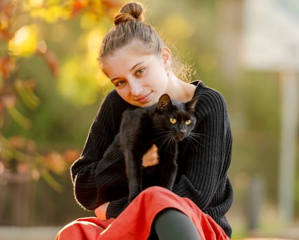 Pretty girl in red skirt hugging black cat outdoors at street with autumn leaves. Beautiful model teenager sitting with feline animal at park