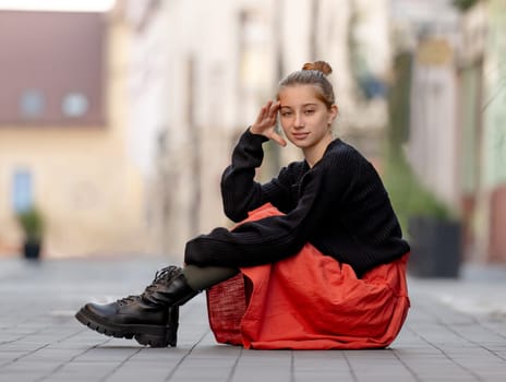 Beautiful teenager girl in red skirt sitting outdoors at street. Pretty teen model posing in trendy clothes