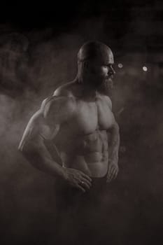 A muscular bald man poses shirtless in the dark amid smoke. Vertical photo