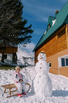 Little smiling girl sits on a sled near a snowman near a wooden chalet. High quality photo