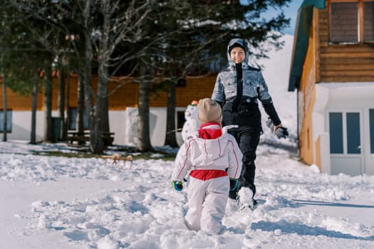 Small child goes to his mother standing next to a snowman in the yard. Back view. High quality photo