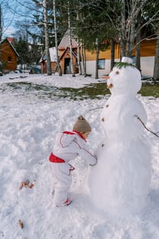 Little girl decorates a snowman with buttons near a wooden house in the snow. Side view. High quality photo