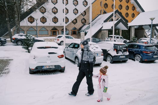 Mother and a little girl walk holding hands past cars parked outside a snow-covered hotel. Side view. High quality photo