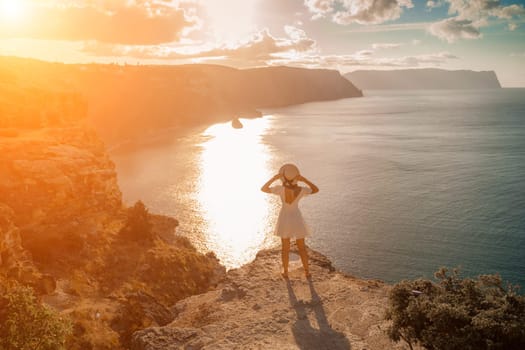 woman sea travel. photo happy woman with hat in white dress standing on the shore on a hill overlooking the sea. golden hour, silhouette of a woman at sunset on the mountain
