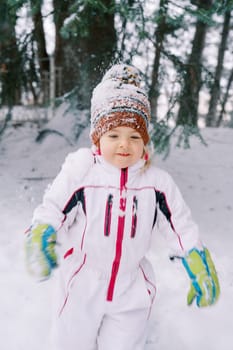 Little girl in a ski suit and a snowy hat walks through the winter forest. High quality photo