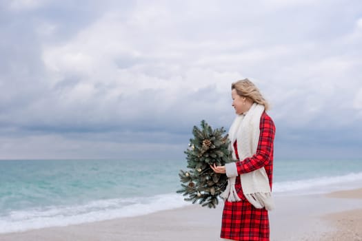 Blond woman holding Christmas tree by the sea. Christmas portrait of a happy woman walking along the beach and holding a Christmas tree in her hands. Dressed in a red coat, white dress