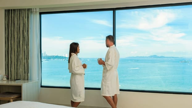 a couple on vacation drinking coffee in the morning looking out the window from their hotel room over the beach and ocean during a luxury holiday