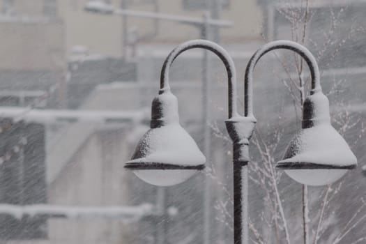 Street lamps covered with snow during a blizzard with blurred buildings in the background.
