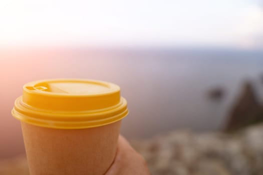 Hand holding Yellow cup with lid, coffee against a backdrop of a blue sky and sea. Illustrating cup and beverage.
