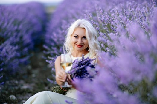 Blonde lavender field holds a glass of white wine in her hands. Happy woman in white dress enjoys lavender field picnic holding a large bouquet of lavender in her hands . Illustrating woman's picnic in a lavender field