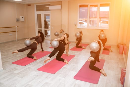 A group of six athletic women doing pilates or yoga on pink mats in front of a window in a beige loft studio interior. Teamwork, good mood and healthy lifestyle concept