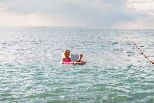 Woman works on laptop in sea. Freelancer, blond woman in sunglases floating on an inflatable big pink donut with a laptop in the sea at sunset. People summer vacation rest lifestyle concept