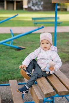 Little girl with a teddy bear sits on a wooden bench in the playground. High quality photo