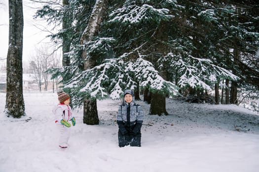 Little girl looks at her smiling mother sitting under a snow-covered spruce tree in a coniferous forest. High quality photo