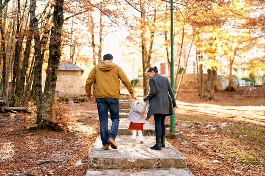 Mom and dad with a little girl walk hand in hand along a paved path in the park, lifting her above the steps. High quality photo