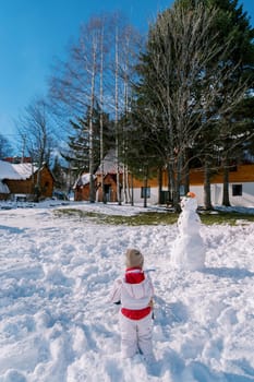 Little girl stands next to a snowman in the courtyard of a wooden house. Back view. High quality photo