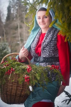 Girl in thick coat and a red sash with basket of fir branches and berries in cold winter day in a forest. Medieval peasant woman with firewood. Photoshoot in stile of Christmas fairy tale