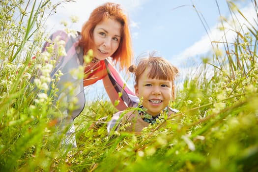 Happy female family with mother and daughter on green and yellow meadow full of grass and flower. Woman with red hair and blonde girl having fun, joy and hug in sunny summer day. Concept family love