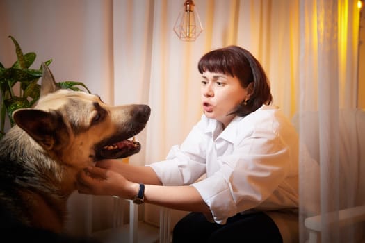 Adult mature woman with big shepherd dog in white shirt. Room with girl and calm cozy evening atmosphere with transparent curtains and soft warm light of lamps. Concept of love for animals, pets