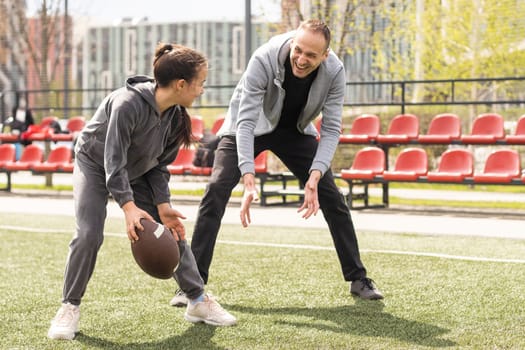 Handsome dad with his little cute daughter are having fun and playing American football on green grassy lawn.