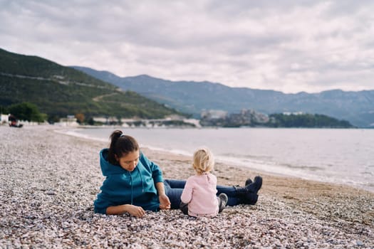 Mom with a little girl are sitting on the beach by the sea and playing with pebbles. High quality photo