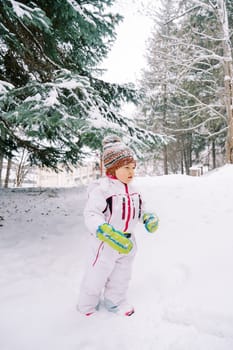 Little girl with snow on her hat stands near a snow-covered fir tree. High quality photo
