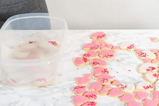 Storing heart-shaped sugar cookies with pink and white royal icing in a large plastic container.