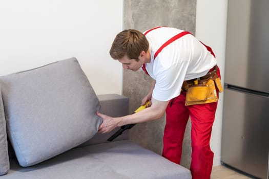 Rear View Of Young Male Worker Cleaning Sofa With Vacuum Cleaner.