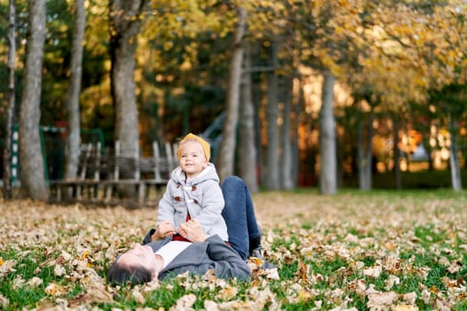 Little smiling girl sits on her mother belly on the autumn lawn, holding her hands. High quality photo