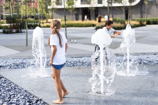 Cheerful young teen girl in city fountain, girl in wet clothes is having fun and enjoying the cool summer water, background city architecture. High quality photo