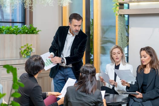 Caucasian manager man stand and hold paper with graph to explain to his colleagues to make the understand of the project and they stay in coworking space.