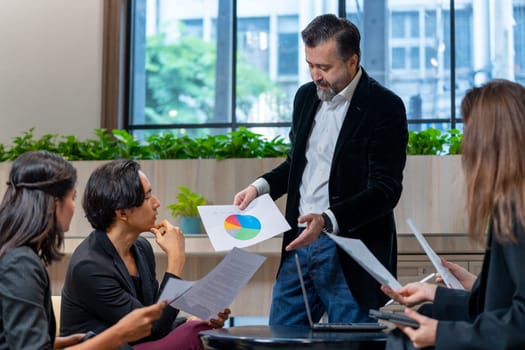 Front view of Caucasian manager man stand and hold paper with graph to explain to his colleagues to make the understand of the project and they stay in coworking space.