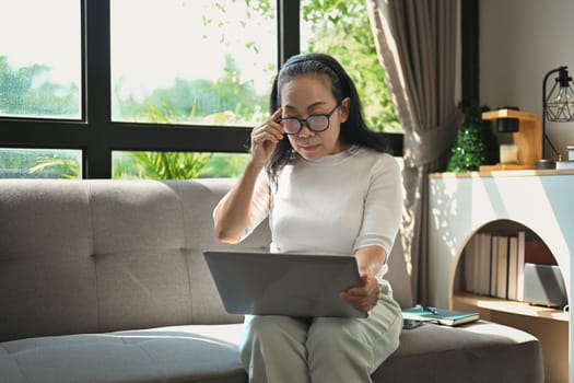Middle age woman touching glasses and reading email on laptop while relaxing on the couch at home