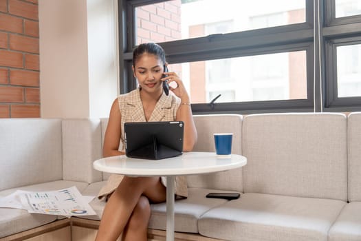 Asian woman use mobile phone during work with tablet to contact her coworker or staff and she sit in area of her office with happiness.