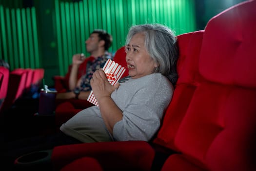 Asian senior woman hold popcorn and look scary during watch movie in cinema theater and Caucasian woman sit near her on the same roll of seats.