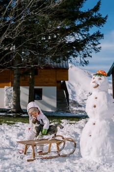 Little girl strokes a striped cat sitting on a sleigh near a snowman in the yard. High quality photo