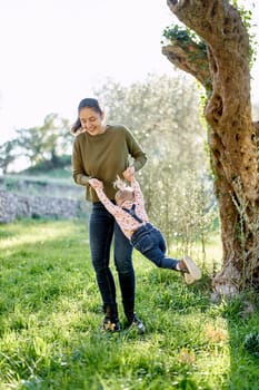 Smiling mother swings a little girl by the arms near a tree. High quality photo