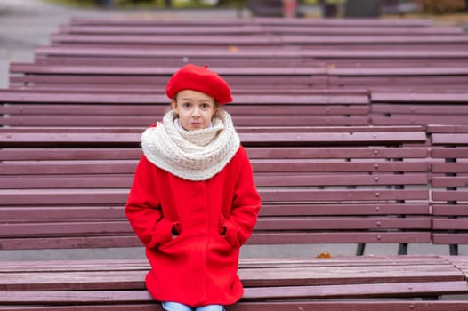 A sad Caucasian girl in a red coat and a beret sits alone on a bench