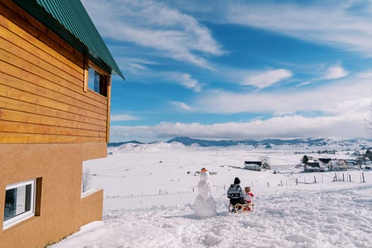 Mom and a small child are sitting on a sleigh near the snowman in front of a wooden house and look at the mountains. Back view. High quality photo