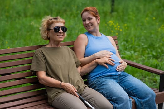 An elderly blind woman holds her pregnant daughter by the belly while sitting on a bench in the park