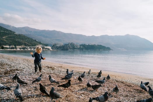 Little girl walks along the seashore with outstretched arms towards a flock of pigeons. High quality photo