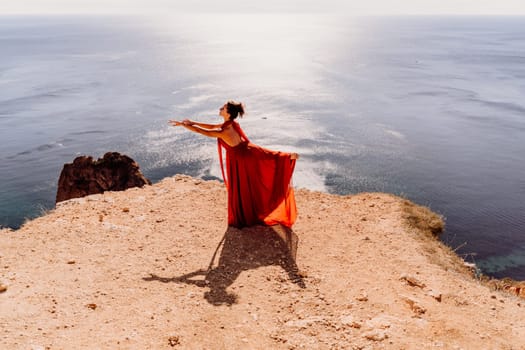 Woman red dress sea. Female dancer posing on a rocky outcrop high above the sea. Girl on the nature on blue sky background. Fashion photo