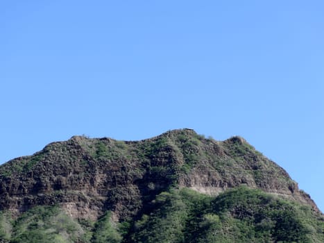 Green crater rim of Diamond Head, a volcanic tuff cone on the island of Oʻahu, Hawaii. The crater is a natural wonder that was formed by a volcanic eruption about 300,000 years ago. The crater is now a state monument and a popular hiking destination. The photo was taken from a distance, so the crater appears small in the frame, but it is actually 3,520 feet in diameter and 761 feet in height. The sky is a clear blue, and the crater contrasts with the surrounding landscape.