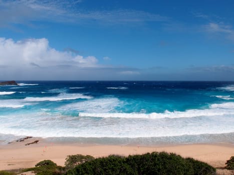 Stunning view of Makapuu Beach and the Pacific Ocean from a high vantage point. Makapuu is a scenic spot on the southeastern tip of Oʻahu, Hawaii. The beach is known for its white sand, turquoise water, and powerful waves. The photo shows the contrast between the sandy shore, the rocky coastline, and the deep blue ocean. The sky is blue with some clouds, adding to the beauty of the scene.